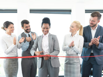 African american woman cutting red ribbon while colleagues are clapping hands at office