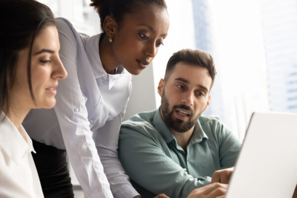 Multiethnic team of focused young managers working on Internet project together, using laptop at office table, discussing online sales strategy, pointing at screen, talking. Vertical shot